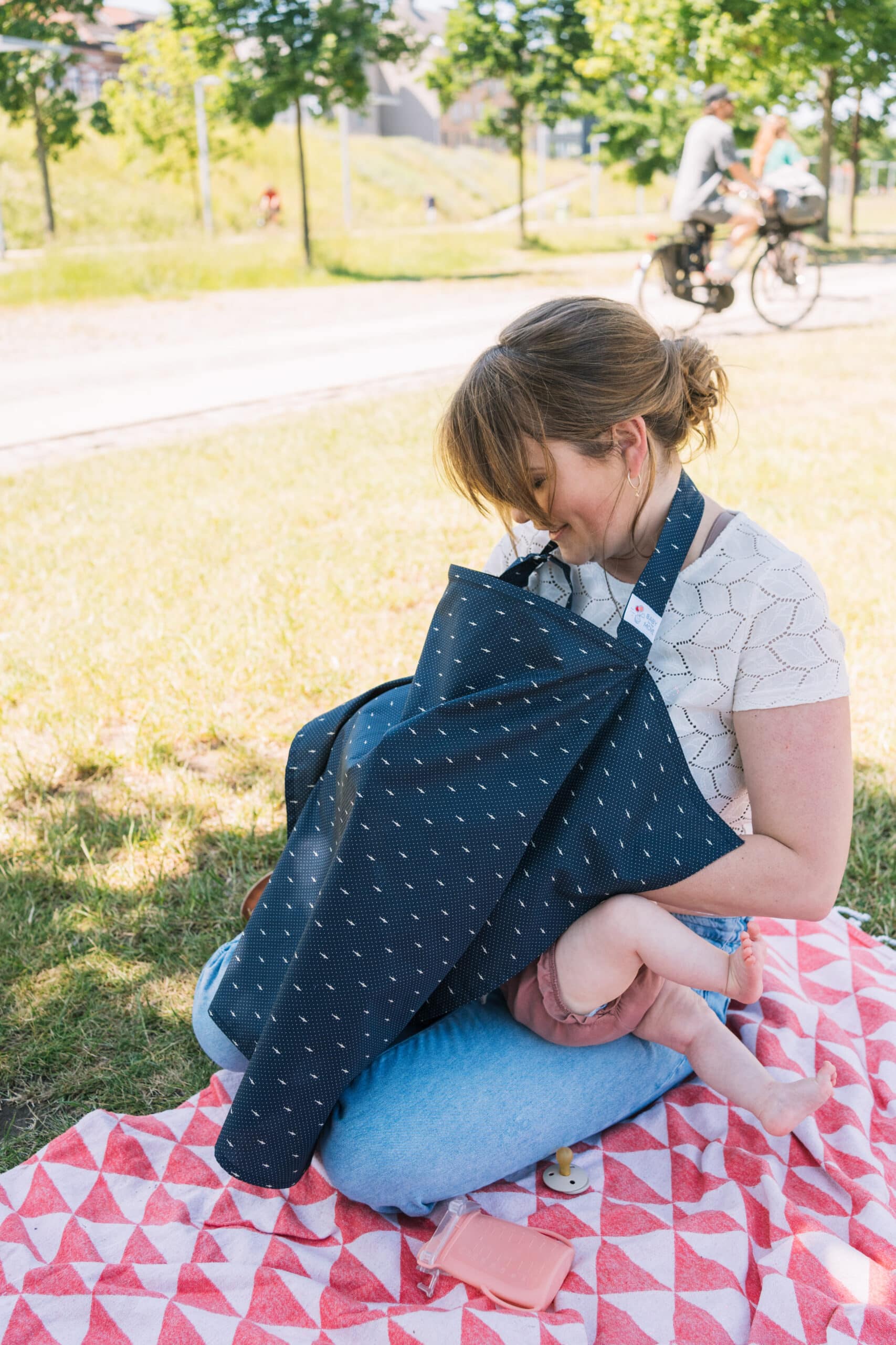 Mom breastfeeding on a picnick blanket in the park wearing our Nursing Cocoon